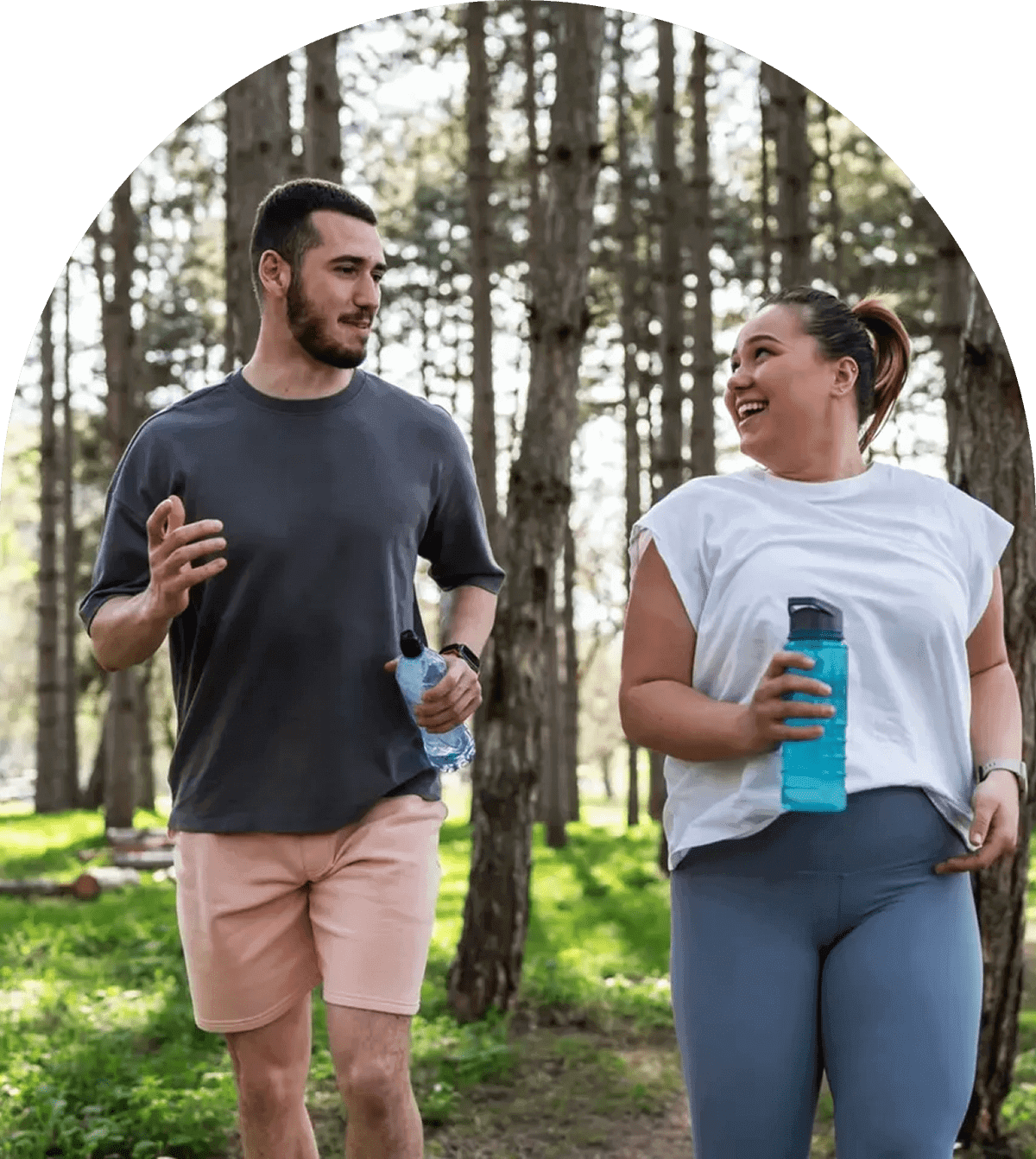 Two friends walk and talk in a sunny forest, holding water bottles.