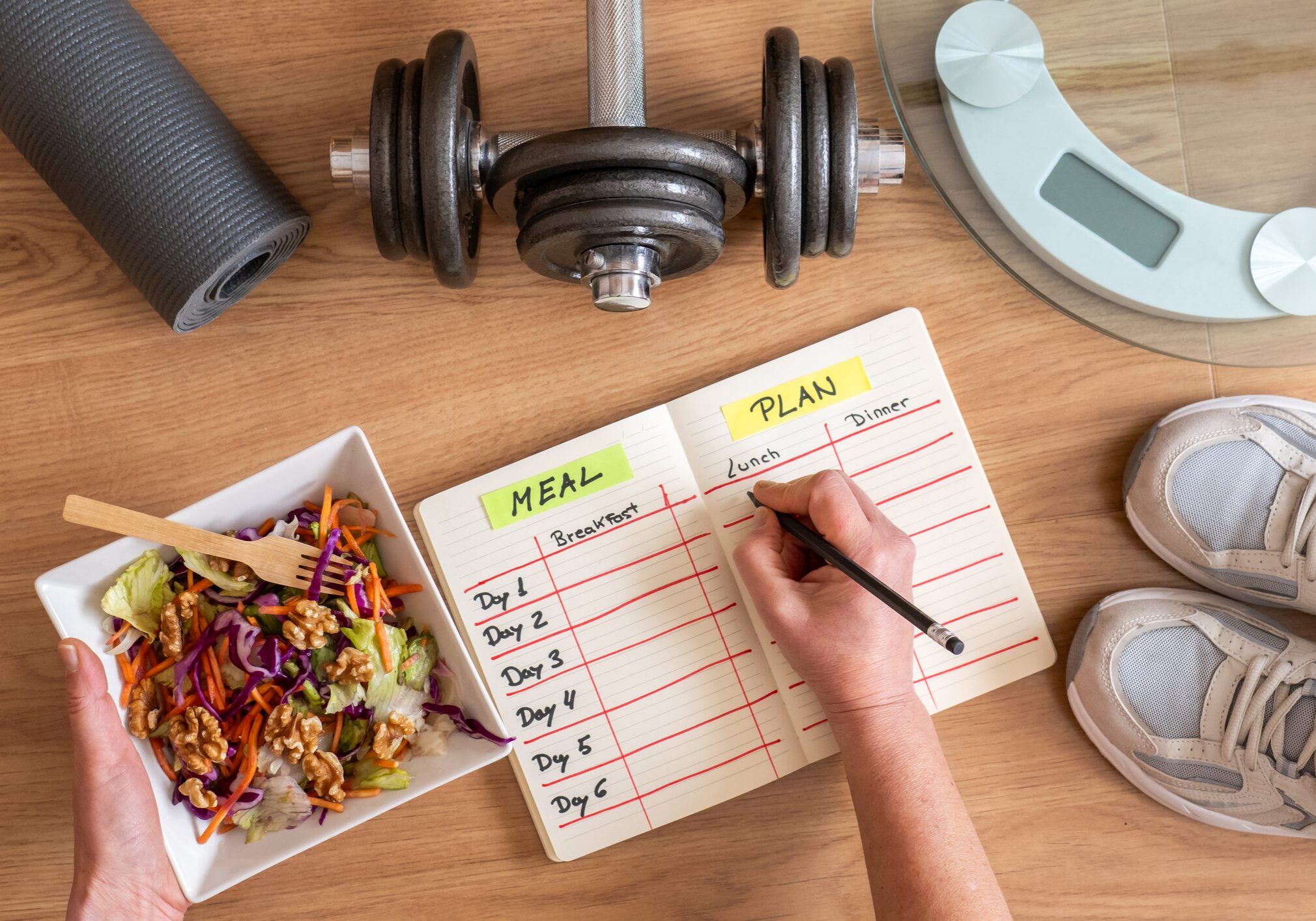 Mature woman writing down her meal plan in a notebook, with a healthy salad, dumbbells, a yoga mat, and a scale on the wooden floor, planning her diet and training schedule
